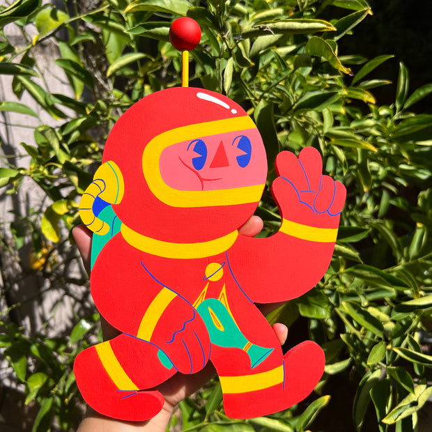 Colorful die cut wooden sculpture of a cartoon character with wearing a red space suit with yellow accents, held against a green leafy background.