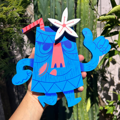 Blue cartoon-like character shaped like a tiki mug with a white flower and straw coming out the top, held by a hand against a green leafy background