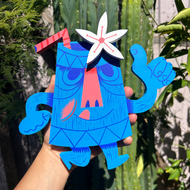 Blue cartoon-like character shaped like a tiki mug with a white flower and straw coming out the top, held by a hand against a green leafy background