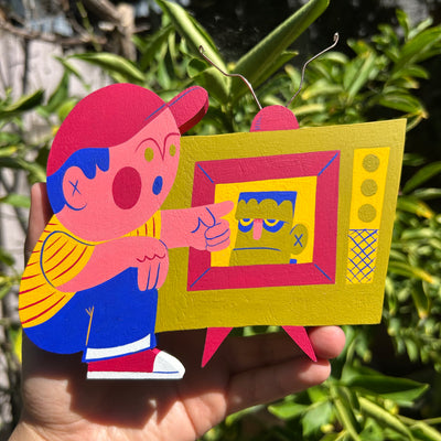 Colorful die cut wooden sculpture of a kid wearing a red baseball cap, striped shirt and jeans and pointing at a retro style TV that plays Frankenstein on it.