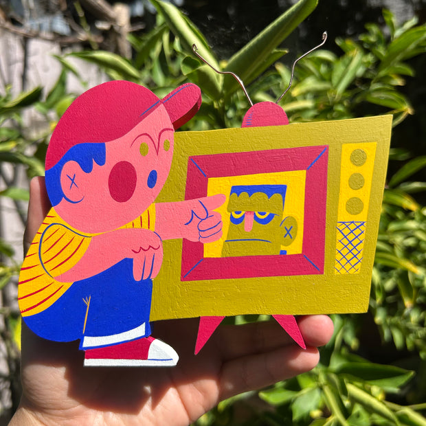 Colorful die cut wooden sculpture of a kid wearing a red baseball cap, striped shirt and jeans and pointing at a retro style TV that plays Frankenstein on it.