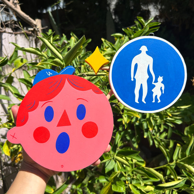 Die cut sculpture of a pink cartoon character's head, wearing a tiny LA cap with a surprised expression. Attached to him is a cross walk sign.