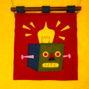 Embroidered wool wall hanging with a design of a cube robot's head with a light coming out the top.