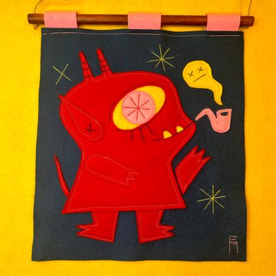 Felt wall hanging with the design of a red devil creature with yellow eyes and pink accents on a dark blue background, hanging against a yellow wall.