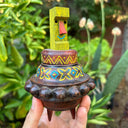 Decorative wooden object, shaped loosely like UFO with a colorful green Tiki head figure, held in front of green foliage