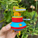 Colorful wooden toy of gator character in a UFO like structure with a laser beam pointing out the front, held in front of green foliage