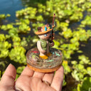 Sculpture of a cat wearing fishing gear and holding a fishing rod with a hooked fish on a base with resin water, fish, rocks and grass.