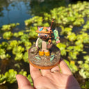 Sculpture of a cat wearing fishing gear and holding a fishing rod with a hooked fish on a base with resin water, fish, rocks and grass.