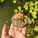 Sculpture of a cat wearing fishing gear and holding a fishing rod with a hooked fish on a base with resin water, fish, rocks and grass.