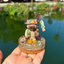Sculpture of a cat wearing fishing gear and holding a fishing rod with a hooked fish on a base with resin water, fish, rocks and grass.