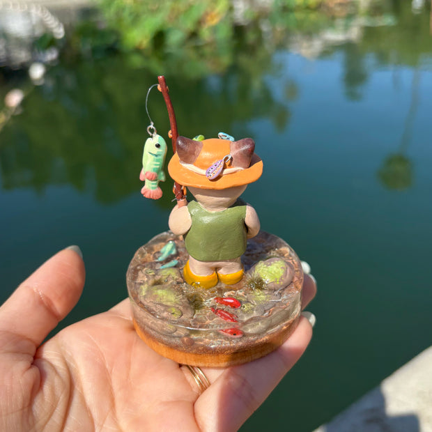 Sculpture of a cat wearing fishing gear and holding a fishing rod with a hooked fish on a base with resin water, fish, rocks and grass.