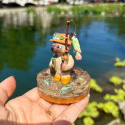 Sculpture of a cat wearing fishing gear and holding a fishing rod with a hooked fish on a base with resin water, fish, rocks and grass.