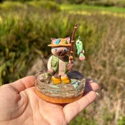 Sculpture of a cat wearing fishing gear and holding a fishing rod with a hooked fish on a base with resin water, fish, rocks and grass.