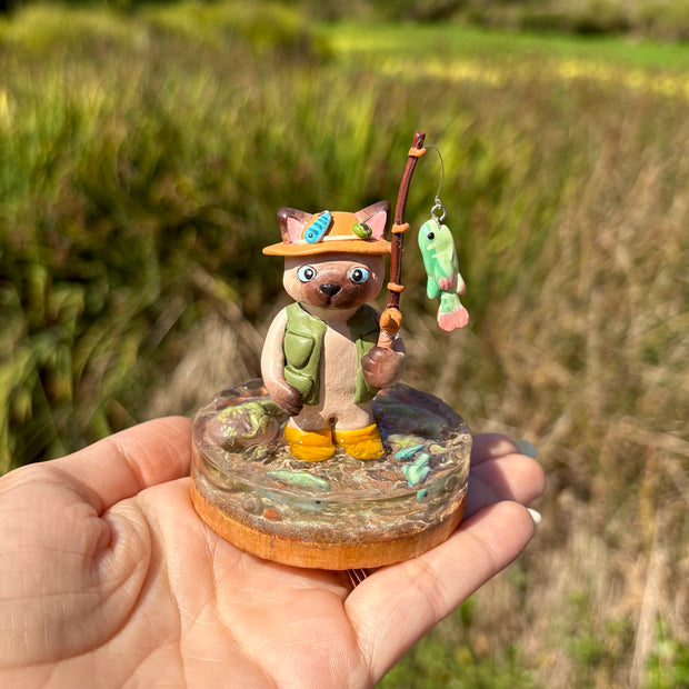 Sculpture of a cat wearing fishing gear and holding a fishing rod with a hooked fish on a base with resin water, fish, rocks and grass.