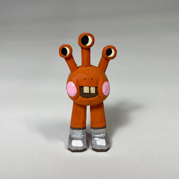 Orange red whittled wooden sculptures of a space alien, with 3 eyes and 2 legs without any other body features. It smiles with buck teeth and wears shiny silver shoes.