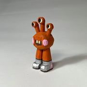 Orange red whittled wooden sculptures of a space alien, with 3 eyes and 2 legs without any other body features. It smiles with buck teeth and wears shiny silver shoes.