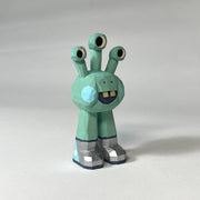 Mint green whittled wooden sculptures of a space alien, with 3 eyes and 2 legs without any other body features. It smiles with buck teeth and wears shiny silver shoes.