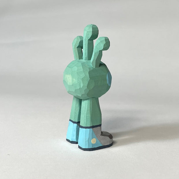 Mint green whittled wooden sculptures of a space alien, with 3 eyes and 2 legs without any other body features. It smiles with buck teeth and wears shiny silver shoes.