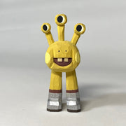 Yellow whittled wooden sculptures of a space alien, with 3 eyes and 2 legs without any other body features. It smiles with buck teeth and wears shiny silver shoes.