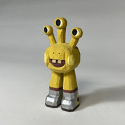 Yellow whittled wooden sculptures of a space alien, with 3 eyes and 2 legs without any other body features. It smiles with buck teeth and wears shiny silver shoes.