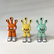 3 painted whittled wooden sculptures of space aliens, with 3 eyes and 2 legs without any other body features. They all smile with buck teeth and wear shiny silver shoes.