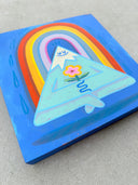 Painting on blue panel of a cartoon style mountain, with a very pointed top and white snow capping it. It has a closed eye cartoon smile and holds a flower, sitting with crossed legs and levitating off the ground. Behind it is a rainbow.