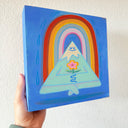 Painting on blue panel of a cartoon style mountain, with a very pointed top and white snow capping it. It has a closed eye cartoon smile and holds a flower, sitting with crossed legs and levitating off the ground. Behind it is a rainbow.