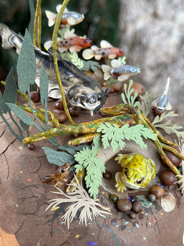 Mixed media diorama of an underwater scene, with a large scary looking fish swimming over a mound of land. Burrowed into the side of the mound is a small frog, hiding from the fish.