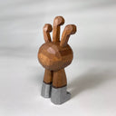 Natural wood whittled wooden sculptures of a space alien, with 3 eyes and 2 legs without any other body features. It smiles with buck teeth and wears shiny silver shoes.