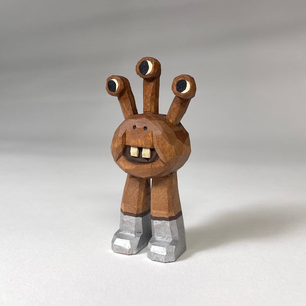 Natural wood whittled wooden sculptures of a space alien, with 3 eyes and 2 legs without any other body features. It smiles with buck teeth and wears shiny silver shoes.
