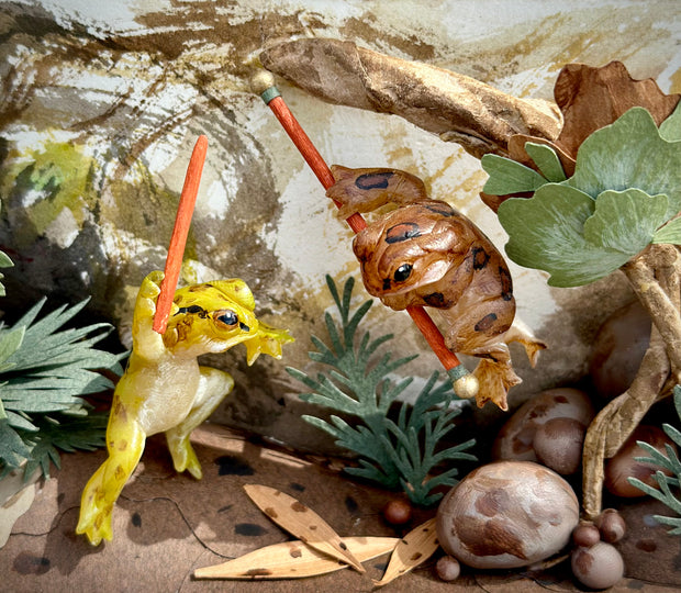 Close up of diorama sculptural work, with 2 small frogs fighting one another, both with staffs. One jumps in the air and the other remains on the ground.