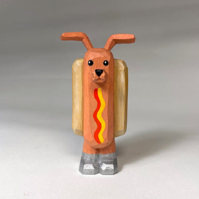 Whittled wooden sculpture of a hot dog, standing up and wearing silver shoes. It has a face of a dog and large protruding dog ears. It has no arms and a squiggle of mustard and ketchup on its front.