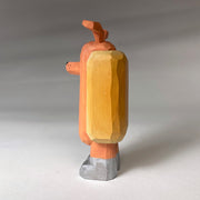 Whittled wooden sculpture of a hot dog, standing up and wearing silver shoes. It has a face of a dog and large protruding dog ears. It has no arms and a squiggle of mustard and ketchup on its front.