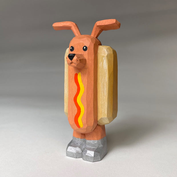 Whittled wooden sculpture of a hot dog, standing up and wearing silver shoes. It has a face of a dog and large protruding dog ears. It has no arms and a squiggle of mustard and ketchup on its front.