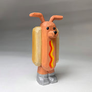 Whittled wooden sculpture of a hot dog, standing up and wearing silver shoes. It has a face of a dog and large protruding dog ears. It has no arms and a squiggle of mustard and ketchup on its front.