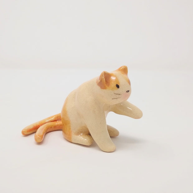 Small ceramic sculpture of a 2-tailed cream colored cat with orange color accents. It sits on its hind legs with one paw extended out. It has a simplistic painted on cat face with whiskers but no mouth.
