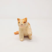 Small ceramic sculpture of a 2-tailed cream colored cat with orange color accents. It sits on its hind legs with one paw extended out. It has a simplistic painted on cat face with whiskers but no mouth.