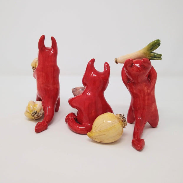  3 red ceramic devil characters with simplistic figures and detailing. One holds up a leek, another holds a cut onion with an onion sitting at its feet. The final one holds a bulb of garlic with another garlic bulb at its feet. Back view.