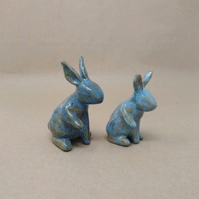 2 small blue ceramic rabbits with brown undertones, they have no defining facial features and sit up with one paw raised.