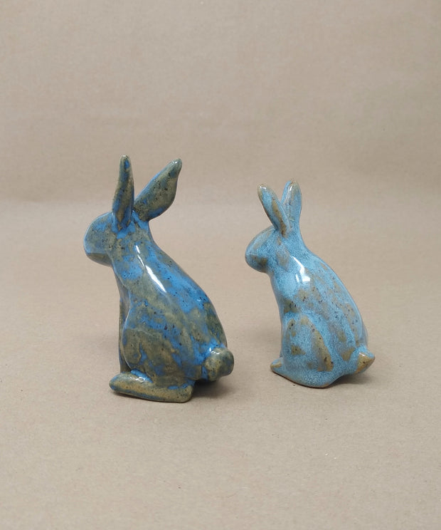 2 small blue ceramic rabbits with brown undertones, they have no defining facial features and sit up with one paw raised.