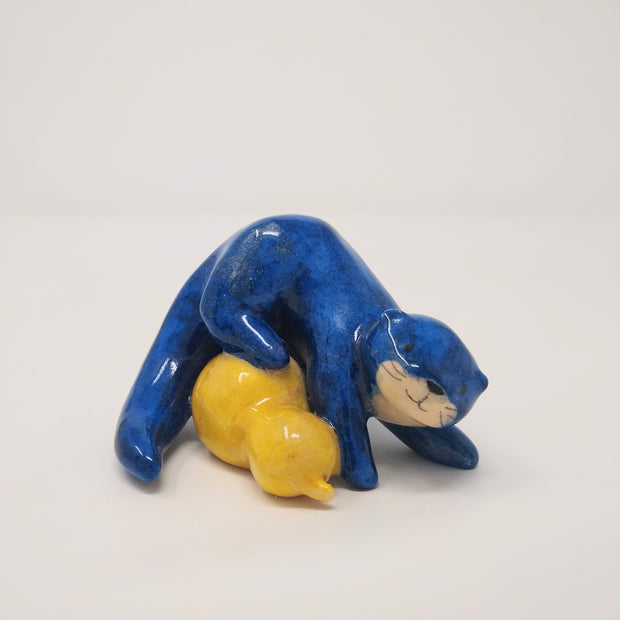 Ceramic sculpture of a blue otter, with its body positioned atop of a yellow gourd. It has a simple smiling face with whiskers.