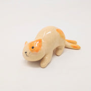 Ceramic sculpture of cream colored with orange spots cat on all fours, leaning with its head tilted as though ready to be petted. It has 2 tails.