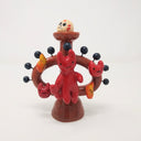 Ceramic sculpture of a small red devil on sitting on a brown vessel, with small blue balls coming out of it and smiling devil faces and flames around it as well. Atop the vessel on a pedestal is a painted skull. 