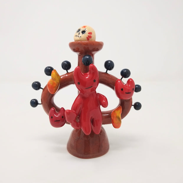 Ceramic sculpture of a small red devil on sitting on a brown vessel, with small blue balls coming out of it and smiling devil faces and flames around it as well. Atop the vessel on a pedestal is a painted skull. 