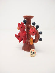 Ceramic sculpture of a small red devil on sitting on a brown vessel, with small blue balls coming out of it and smiling devil faces and flames around it as well. Atop the vessel on a pedestal is a painted skull.