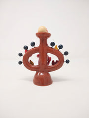 Ceramic sculpture of a small red devil on sitting on a brown vessel, with small blue balls coming out of it and smiling devil faces and flames around it as well. Atop the vessel on a pedestal is a painted skull.