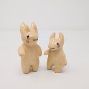 2 small off-white ceramic devil characters, one with 4 horns and the other with only 2. They both have tears but with smiling faces and a hand extended out.