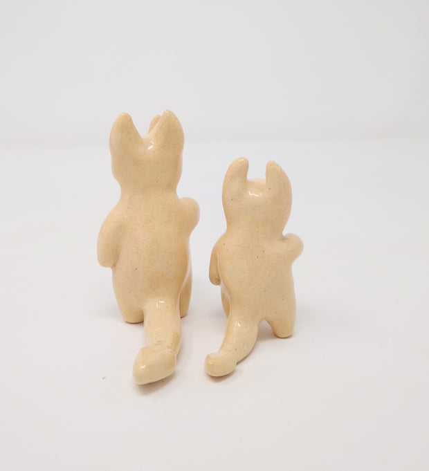 2 small off-white ceramic devil characters, one with 4 horns and the other with only 2. They both have tears but with smiling faces and a hand extended out.