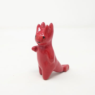 Red ceramic sculpture of a devil like creature, with a smooth body and simple smiling face. It has 4 horns atop its head and one arm extended out, as if waving. 