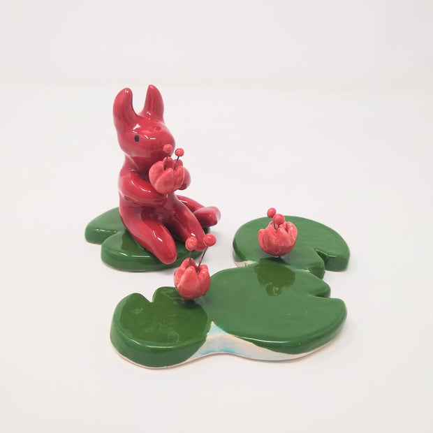 Ceramic sculptures, a cute red devil sits atop of a green lily pad and holds a flower in its hands. Across, is a set of 3 lily pads with flowers blooming out from them.